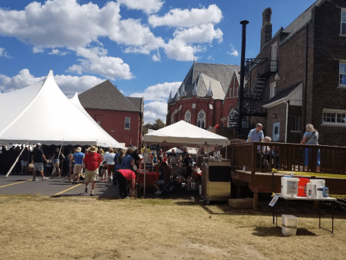 A lively outdoor event with tents, people mingling, and a church in the background under a partly cloudy sky.