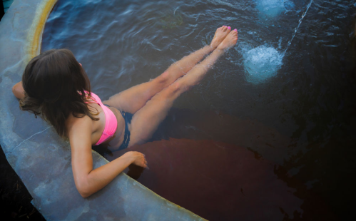 A person relaxes in a circular pool, with their feet submerged in the water and a fountain feature in the background.