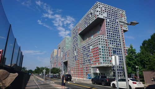 Modern building with a colorful, grid-like facade under a clear blue sky, surrounded by trees and a road.