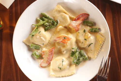 A plate of ravioli topped with lobster, asparagus, and a creamy sauce, served on a wooden table.