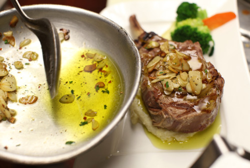 A plated dish featuring a juicy chop topped with garlic and herbs, served with broccoli and a drizzle of olive oil.