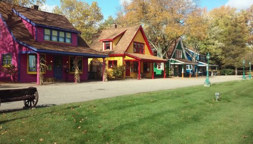 Colorful houses line a gravel path, surrounded by autumn trees and a grassy area.