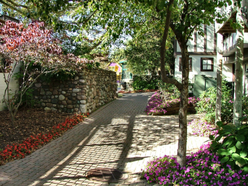 A charming pathway lined with colorful flowers and trees, leading past a stone wall and quaint buildings.