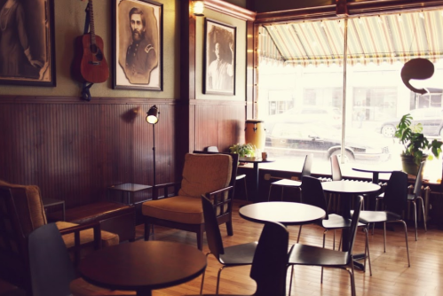 Cozy café interior with wooden walls, vintage portraits, and a mix of chairs and tables by a large window.