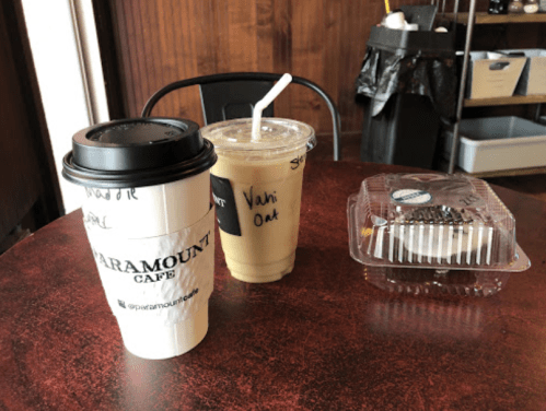 A table with a coffee cup, iced drink, and a dessert in a takeout container at a café.