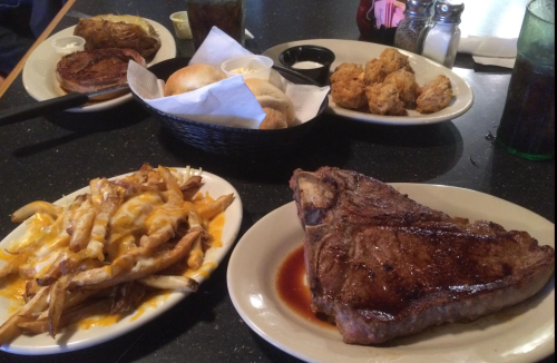 A table spread featuring a large steak, loaded fries, rolls, and sides, with drinks in the background.