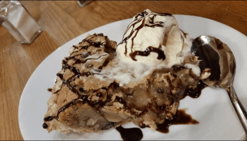 A slice of warm chocolate chip cookie pie topped with vanilla ice cream and drizzled with chocolate sauce on a plate.