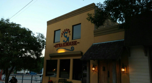 Exterior of a steakhouse building with a colorful sign, surrounded by trees and a clear sky.