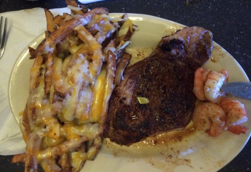 A plate featuring a grilled steak, cheesy fries, and shrimp, served on a white dish with a napkin on the side.