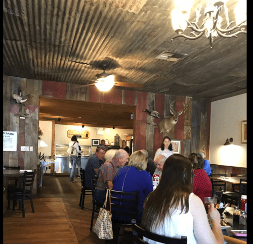 A rustic restaurant interior with wooden walls, diners at tables, and a woman walking towards the kitchen area.