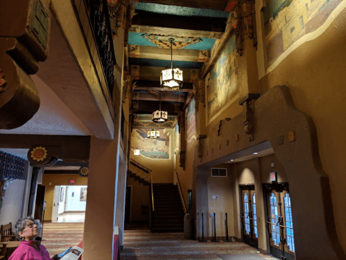 Interior of a building featuring colorful murals, wooden beams, and a staircase, with a person seated nearby.