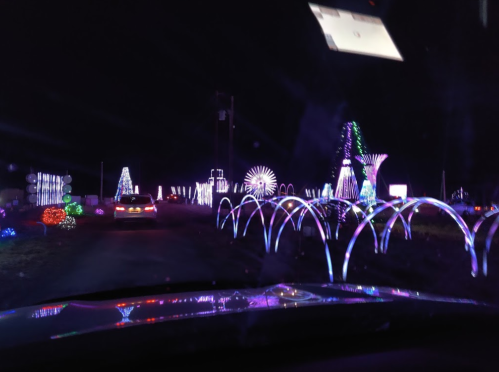 A car drives through a colorful display of illuminated decorations at night, featuring various light structures.