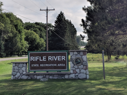 Sign for Rifle River State Recreation Area, surrounded by trees and grass, with a road visible in the background.