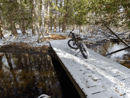 A bike rests on a snowy wooden bridge over a small stream, surrounded by trees in a winter landscape.