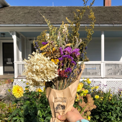 A hand holds a bouquet of dried flowers wrapped in paper, with a house and garden in the background.