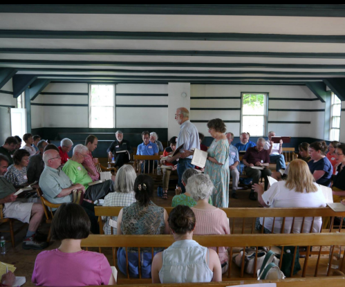A group of people seated in a rustic hall, engaged in a meeting or gathering, with some standing and holding books.