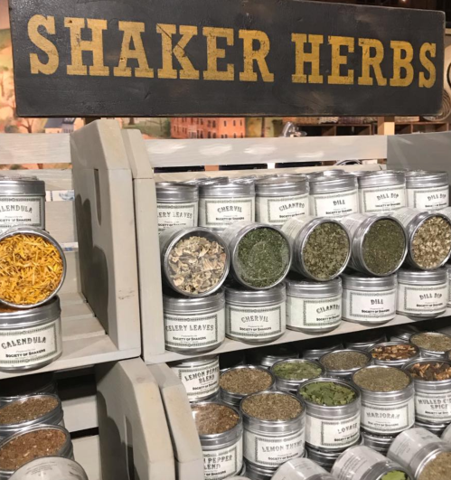 Display of various Shaker herbs in labeled jars on a wooden shelf, with a sign above reading "Shaker Herbs."