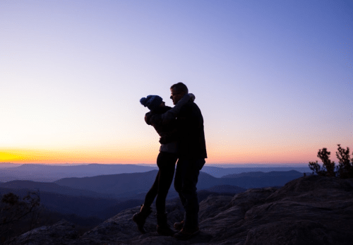A couple embraces on a rocky outcrop at sunset, with mountains and a colorful sky in the background.