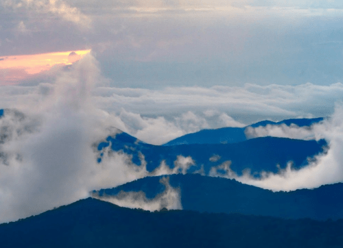 A scenic view of rolling mountains shrouded in clouds, with a hint of sunset peeking through the mist.