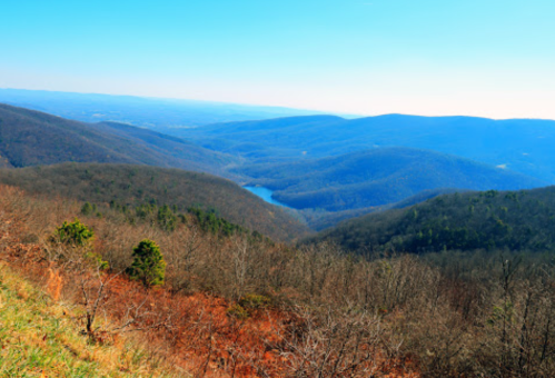 A panoramic view of rolling mountains and a serene lake under a clear blue sky.