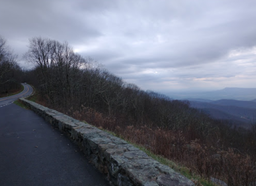 A winding road alongside a stone wall, with a cloudy sky and distant mountains in the background.