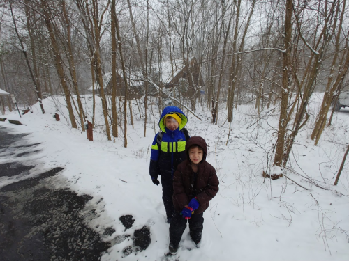 Two children stand in the snow, surrounded by trees, wearing colorful winter jackets and smiling at the camera.