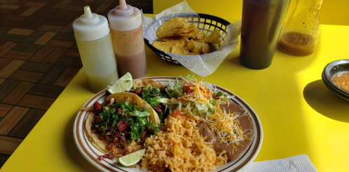 A colorful plate of tacos with rice, beans, and salad, accompanied by sauces and tortilla chips on a bright yellow table.