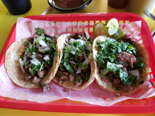 Three tacos filled with meat, onions, and cilantro, served on a red tray with lime wedges.