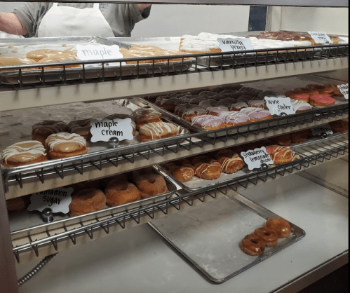 A display case filled with various donuts, including maple, vanilla, and cream varieties, labeled on small signs.