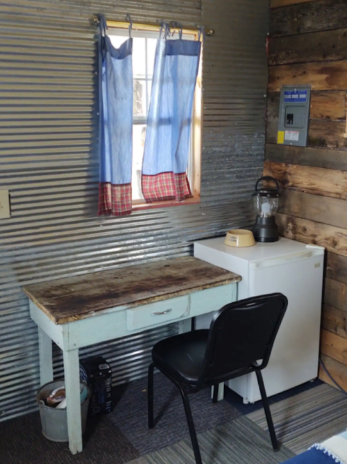 A small room with a wooden desk, black chair, fridge, and a window with blue curtains. Walls are corrugated metal and wood.