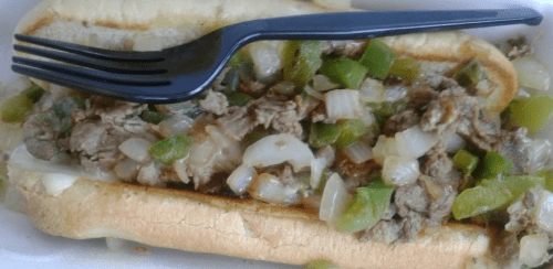 A loaded sandwich with chopped meat, onions, and green peppers, topped with a black plastic fork.