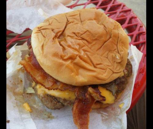 A close-up of a cheeseburger with bacon, nestled in crumpled paper on a red table.