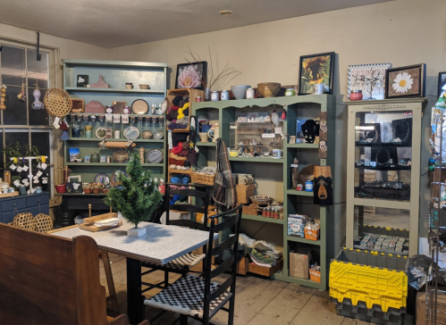 A cozy shop interior with shelves displaying crafts, pottery, and handmade items, featuring a table and chairs.
