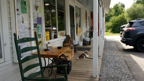 A porch with a green rocking chair, a table, and a dog lying down, beside a gravel path and parked cars.