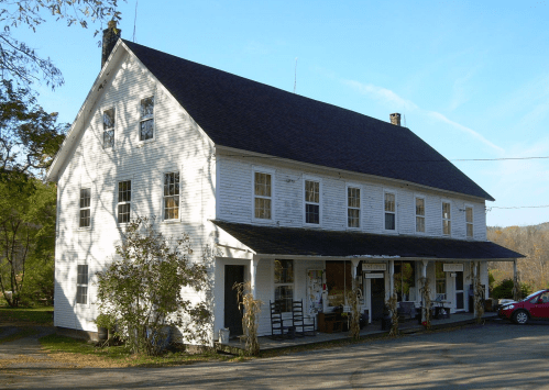A large, white two-story building with a sloped roof, featuring a porch and surrounded by trees and a clear blue sky.