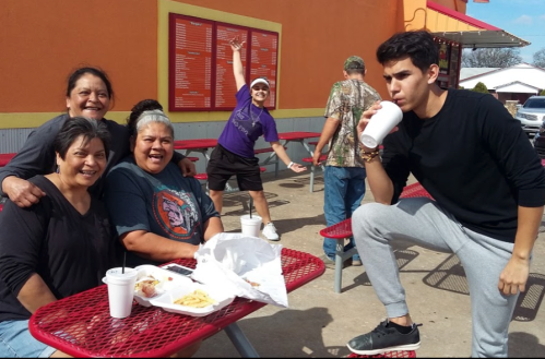 A group of five people enjoying food at a picnic table outside, with one person playfully posing in the background.