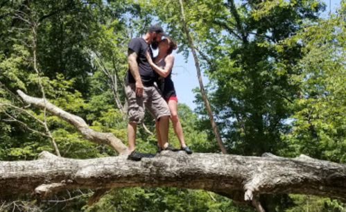 A couple shares a kiss while standing on a large fallen tree in a lush green forest.