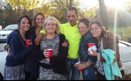 A group of six people, smiling and holding red cups, pose together outdoors with trees in the background.