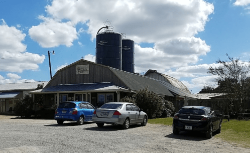 A rustic building with a metal roof, surrounded by cars and silos, under a blue sky with fluffy clouds.
