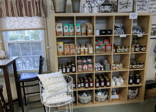 A wooden shelf displays various jars, bags, and boxes of food products, with a chair beside it in a cozy shop setting.