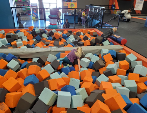 Children play in a colorful foam pit filled with orange, blue, and black blocks at a recreational facility.