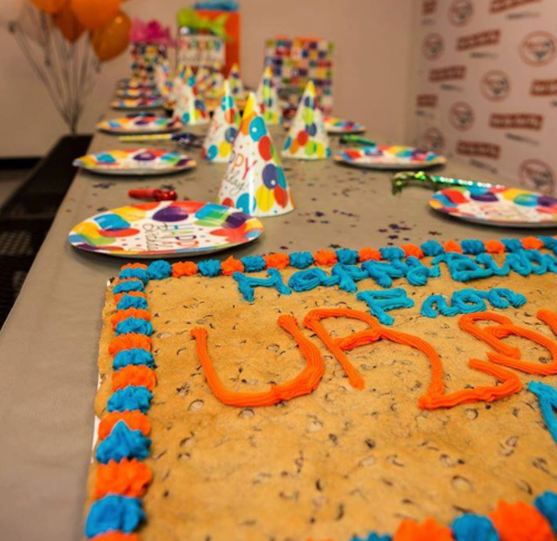 A festive table set for a celebration, featuring a large cookie cake and colorful party hats and decorations.