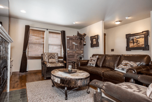 Cozy living room with brown leather sofa, patterned chairs, wooden coffee table, and rustic decor. Natural light from windows.