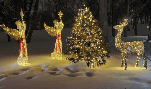 Two illuminated angels and a glowing reindeer beside a decorated Christmas tree in a snowy landscape at night.