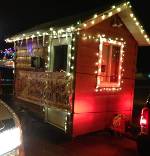 A small, decorated trailer with holiday lights and a sign for "Holiday Lights at Lindenwood Park."