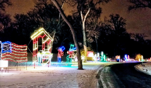 Colorful holiday lights decorate a snowy path, featuring festive displays and trees against a night sky.