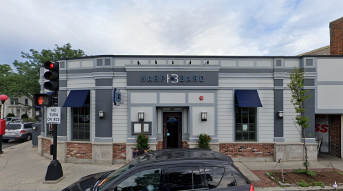 Exterior of a restaurant named "Harp & Bard," featuring blue awnings and a brick facade, with traffic lights nearby.