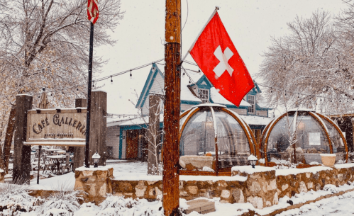 A cozy café with outdoor seating under glass domes, surrounded by snow and a Swiss flag, creating a winter atmosphere.