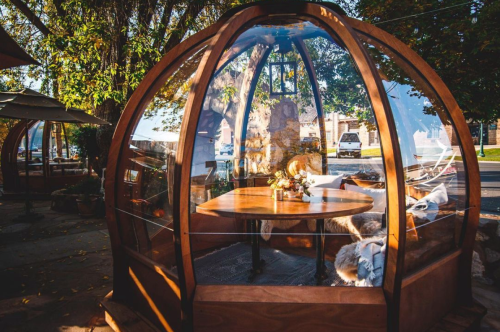A cozy glass dome structure with a wooden frame, featuring a table and seating, surrounded by trees and outdoor scenery.