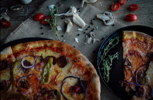 A close-up of a pizza topped with vegetables and herbs, surrounded by cherry tomatoes and mushrooms on a rustic table.